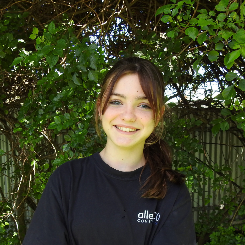 Teen girl with brown hair in a ponytail and long fringe. She is smiling at the camera and is wearing a navy blue t-shirt