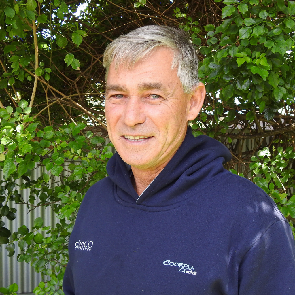 A man with grey hair and smiling at the camera. He is wearing a navy blue hooded jumper