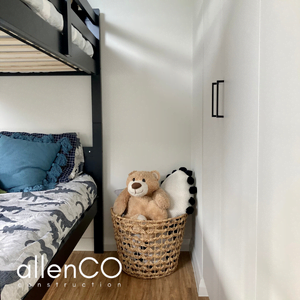 A teddy bear is sitting in a woven basket between a black bunk bed and grey cupboards with black handles