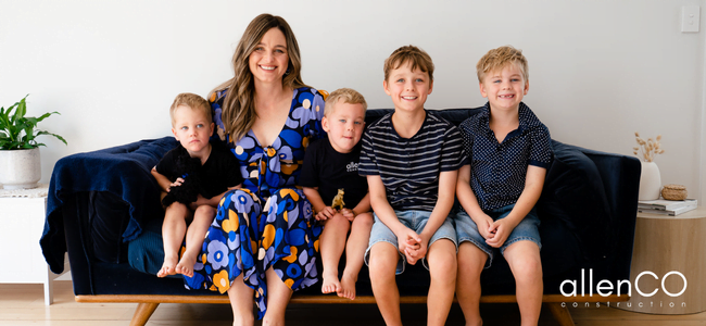 A young woman is sitting on a big blue couch with her four young boys. They are all smiling.