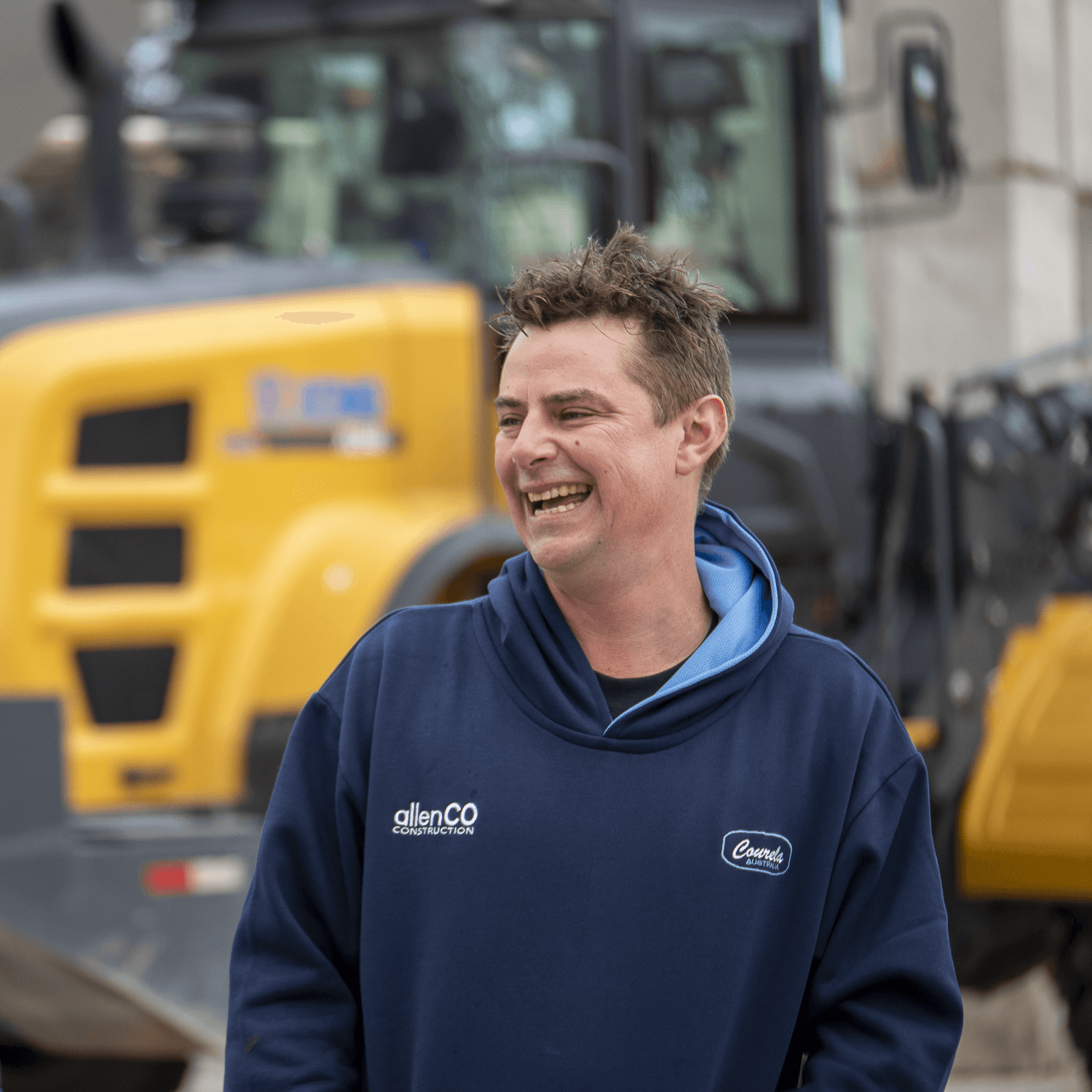 Cameron from allenCO Construction is standing outside and laughing at someone off-camera. There is construction machinery behind him. He is wearing a dark blue hoodie jumper that has a small white logo for allenCO Construction.