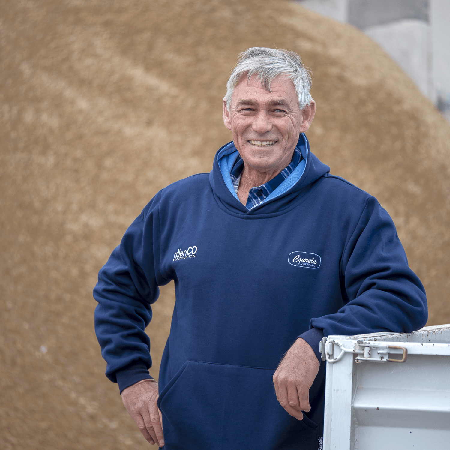 Owner of allenCO Construction, Mark Allen, is smiling at the camera. He is leaning against the tray of a white ute. He is wearing a navy blue hooded jumper that has a small white logo for allenCO Construction on it. There is a pile of construction rubble behind him.