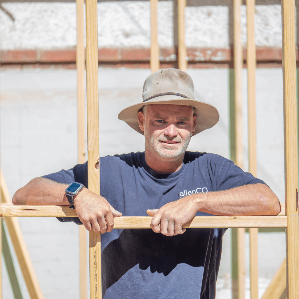 A man wearing a wide brim hat is looking at the camera. He is standing outside in a construction site. He is wearing a navy blue top with the company logo for allenCO Construction.