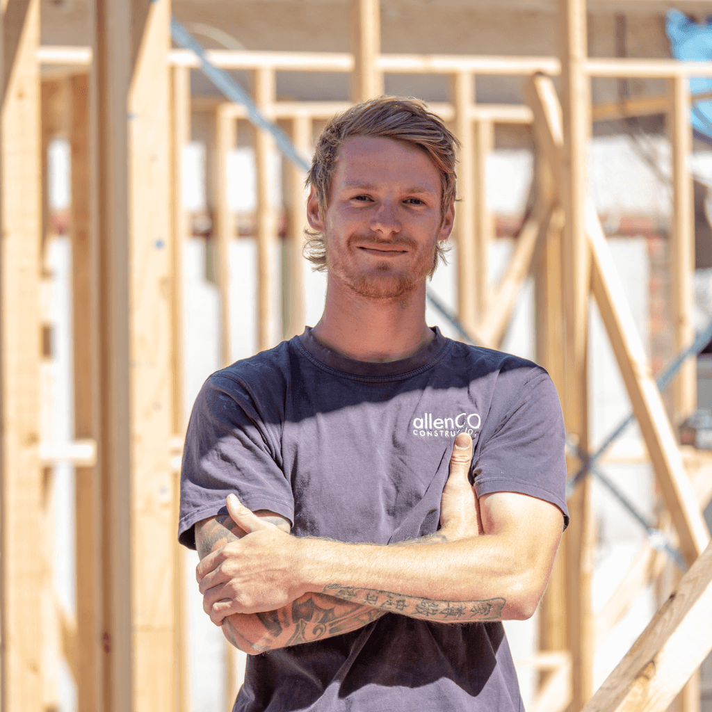 A man has his arms crossed across his body and is looking at the camera smiling. He is standing outside in a construction site. He is wearing a navy blue top with the company logo for allenCO Construction.